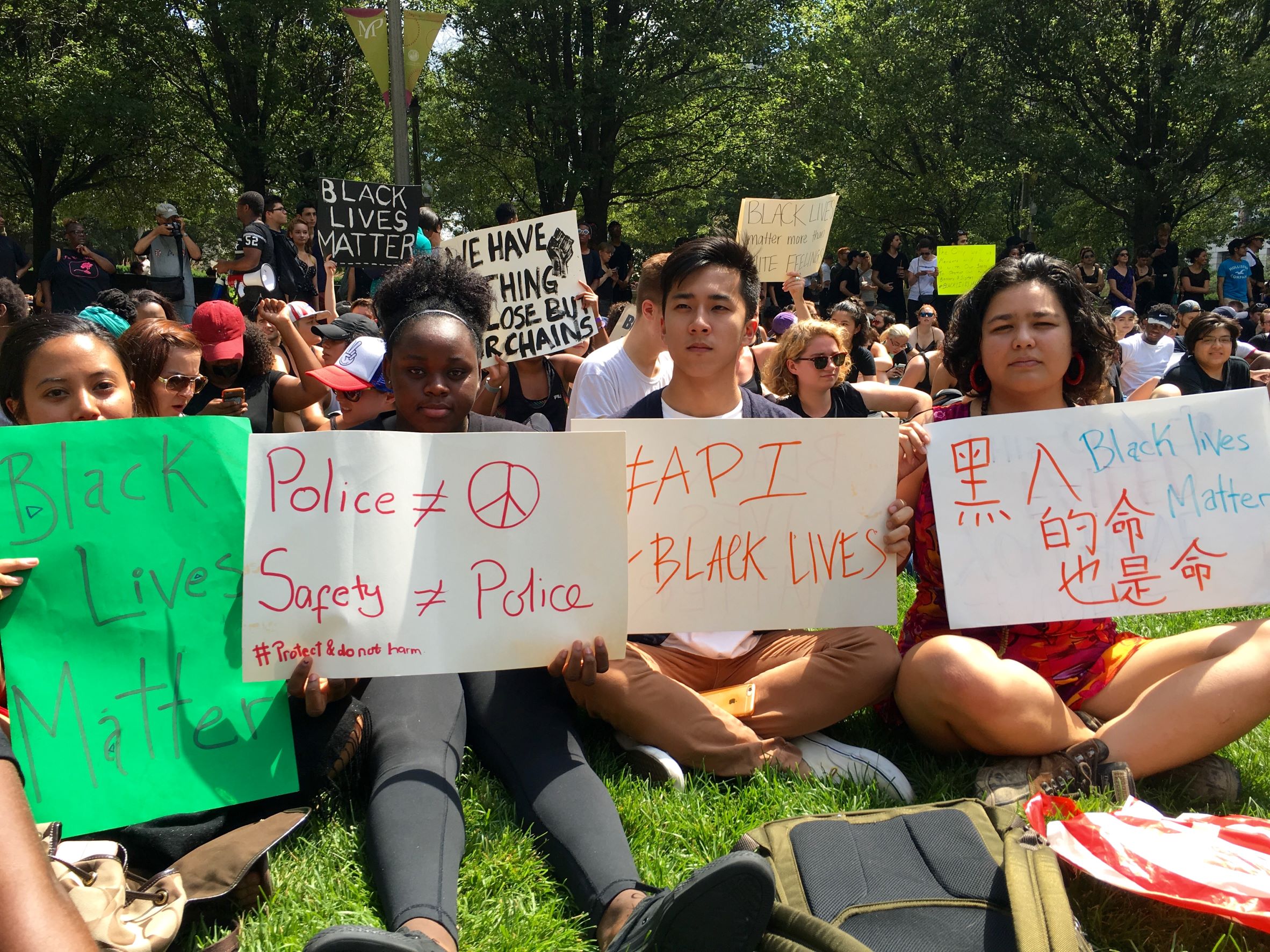 2016 SOL/PEP student Lance Tran sits with a crowd at a Black Lives Matter protest, holding up signs with Black Lives Matter slogans including #API4BlackLives.