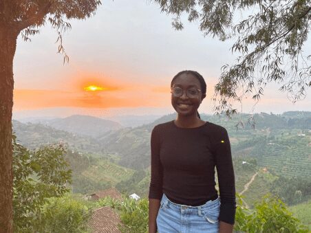 Hart Fellow 2021-22 Kemunto Okindo smiles at the camera. Behind her, a sunset over the hills of Rwanda is visible.