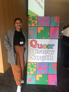 2023-2024 Hart Fellow Sophia Chimbanda standing with a banner that reads "Queer History South"